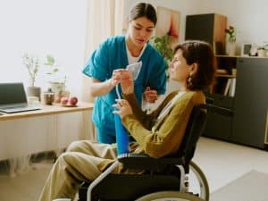 Health provider assist NDIS participant in wheelchair by demonstrating breathing aid usage at home.