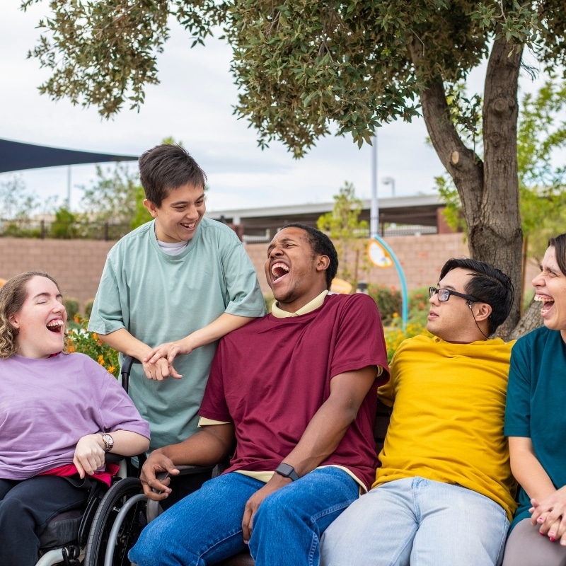 Circle of Support showing diverse adults with disabilities laughing together on an outdoor bench in a community setting